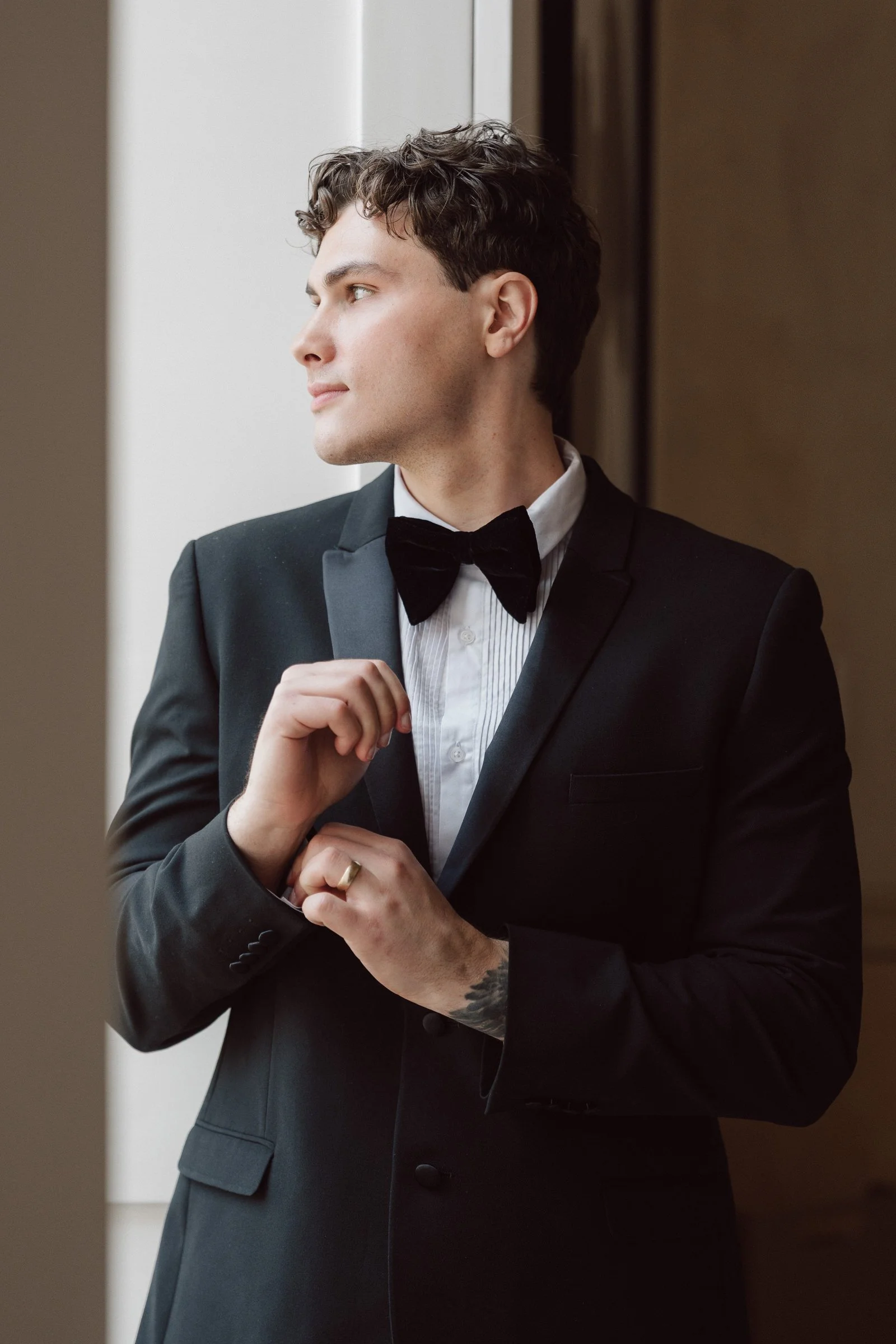 A man in a black tuxedo and bowtie adjusting his cufflink, standing near a wall, looking to his left with a thoughtful expression.