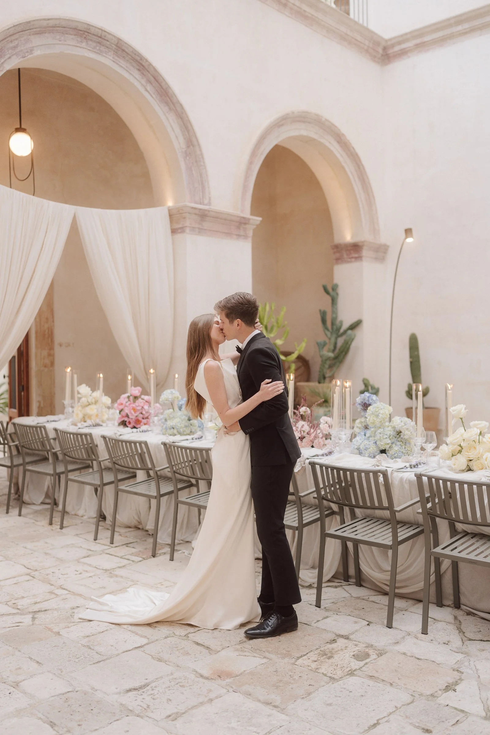 A bride and groom are dancing in a decorated banquet hall with high ceilings, elegant arches, and draped curtains, surrounded by a long table filled with pastel-colored flower arrangements and lit candles.