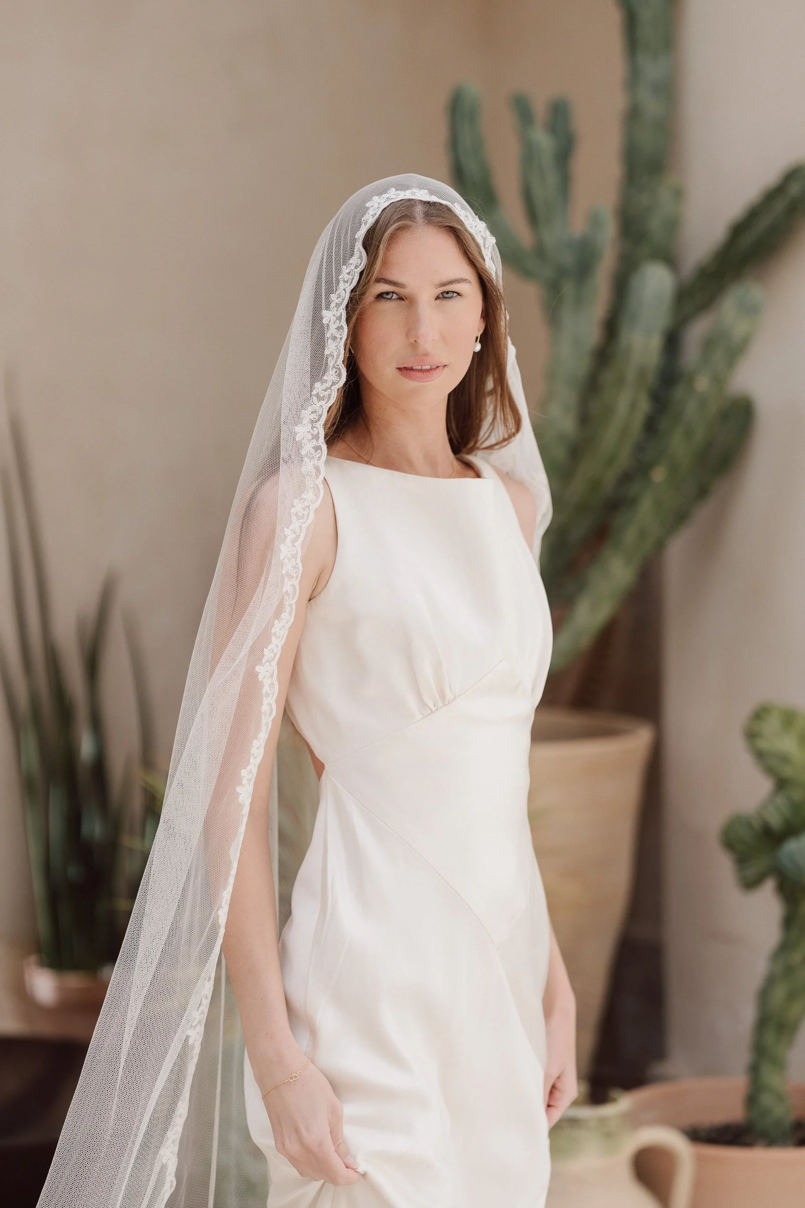 A woman in a white dress and veil standing indoors next to a large cactus plant.