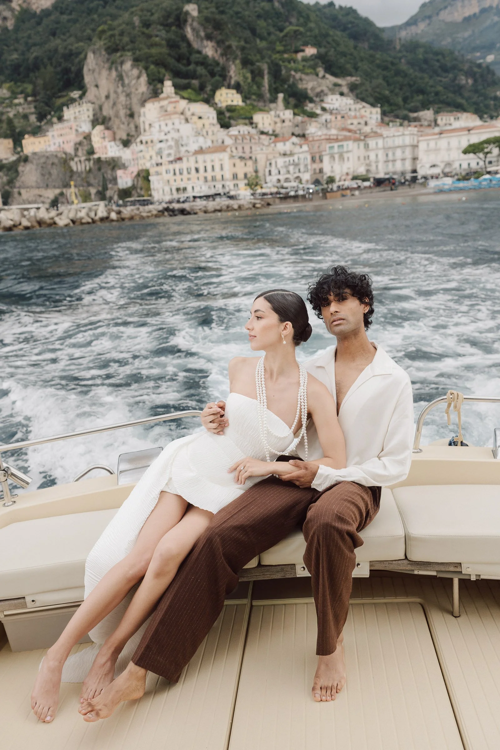A bride and groom sitting on a boat with a coastal town and hillside in the background.