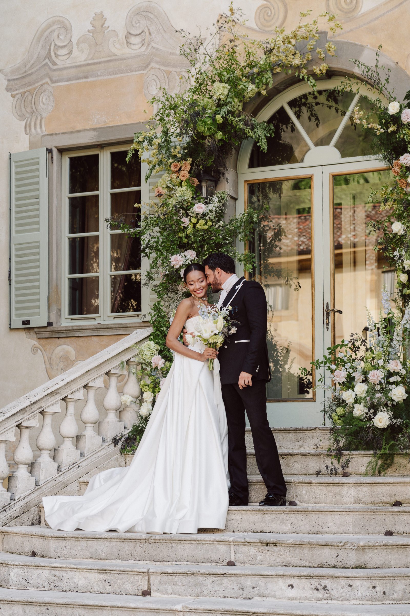 A newlywed couple standing on the steps of a historic building with white trim, surrounded by floral decorations, smiling and sharing an intimate moment.