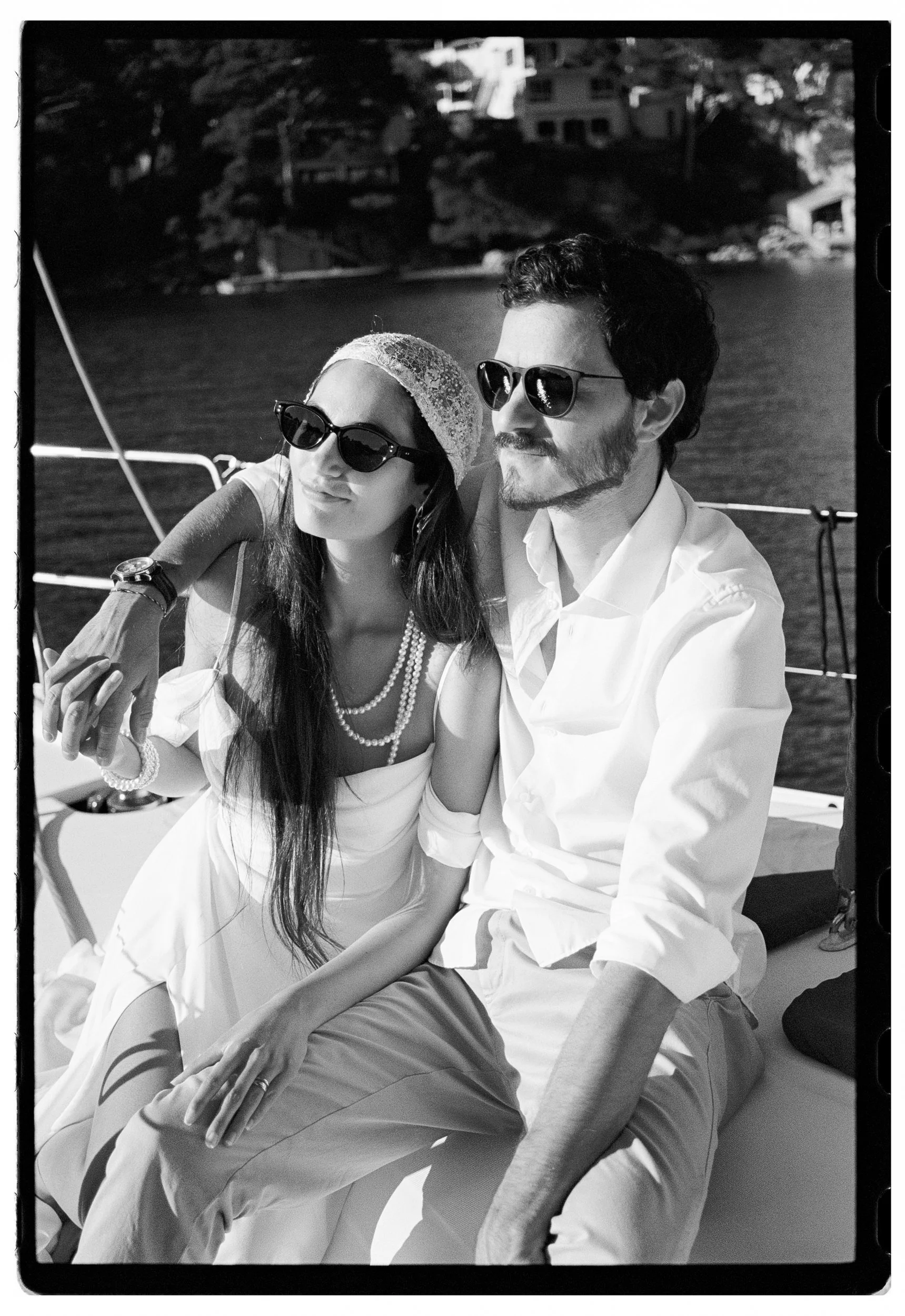 A black and white photo of a man and woman sitting closely together on a boat, with water and house buildings in the background. They are both wearing sunglasses and have relaxed expressions.