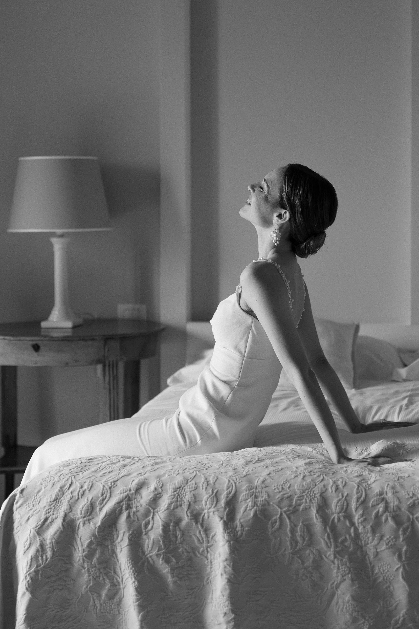 A woman in a wedding dress sitting on a bed with her back arched, smiling with her eyes closed, in a bedroom with a bedside lamp and a table.
