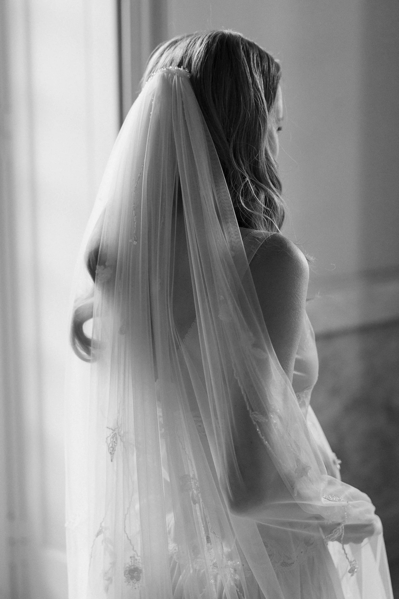 A bride in profile, wearing a wedding dress and veil, looking out a window, in black and white.