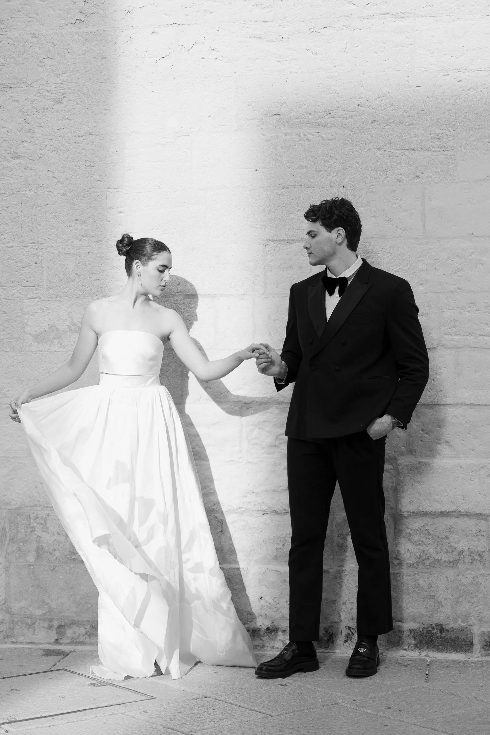 A black and white photo of a young woman wearing a strapless white gown and a young man dressed in a tuxedo with a bow tie. They are holding hands against a textured wall, with their eyes closed and a calm expression.