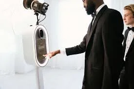 A man in a tuxedo and woman in formal attire using a touchless hand sanitizer dispenser that is mounted on the wall.