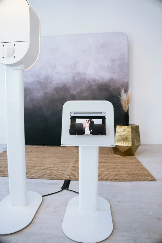 A photobooth displaying a couple kissing, set up in a modern, minimalistic room with a black and white gradient backdrop, a beige rug, and a textured gold vase with dried plants.