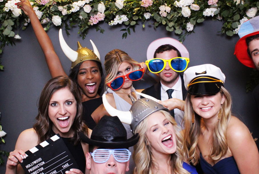 Group of women smiling and taking a photo at a social event, holding glasses of wine or champagne, with a lush green leafy wall and warm wooden interior.