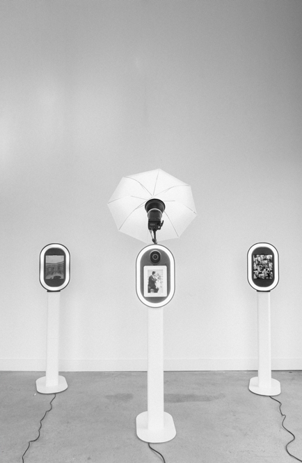 Three digital display machines in an art gallery, with an umbrella hanging above the center display, which is connected to a light fixture.