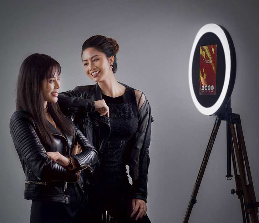 Two women stand chatting in front of a professional ring light with a digital screen. One woman is smiling and wearing a black lace top, the other is looking and wearing a black leather jacket.