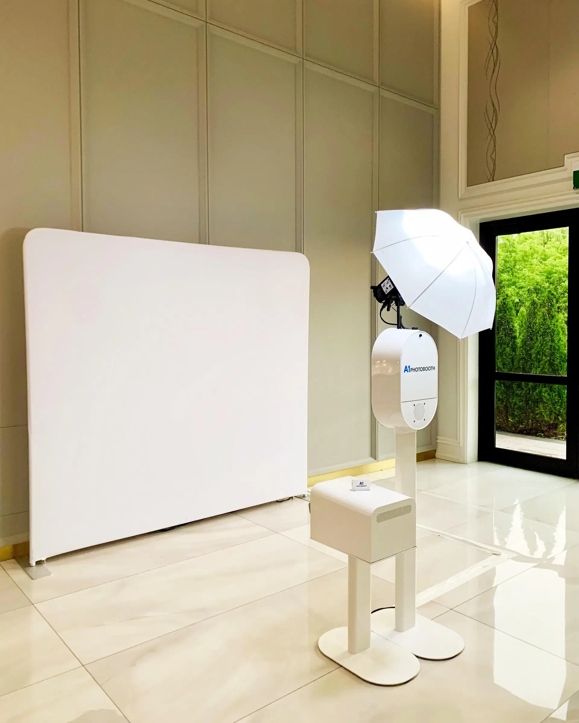 Photo booth setup with white backdrop, umbrella light, and photo printer in a bright room with glossy tile floor and glass door.