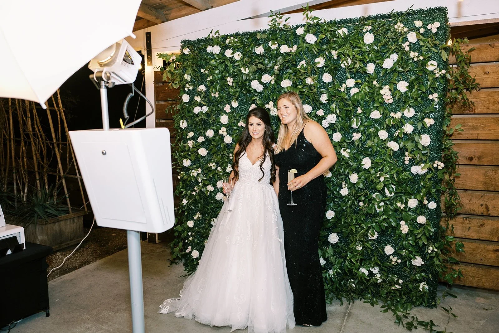 Group of women smiling and taking a photo at a social event, holding glasses of wine or champagne, with a lush green leafy wall and warm wooden interior.