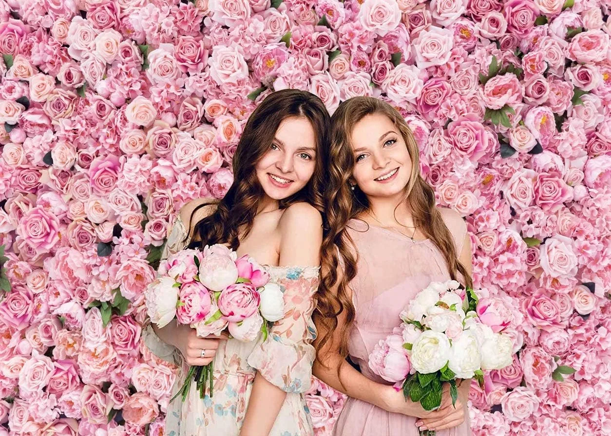 Two young women with long wavy hair smiling and holding bouquets of pink and white flowers, standing in front of a wall covered with pink roses and peonies.