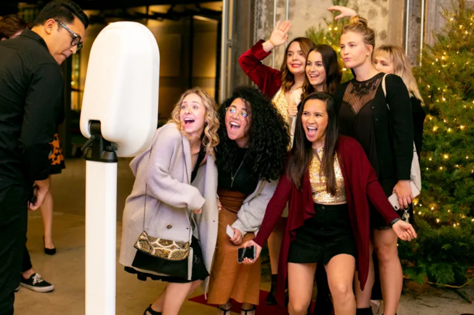 Five friends at a celebration, dressed elegantly, holding drinks, with a floral backdrop. The women wear flowy dresses and wide-brimmed hats with flowers; one woman has large earrings. The man wears a light suit with a boutonniere.