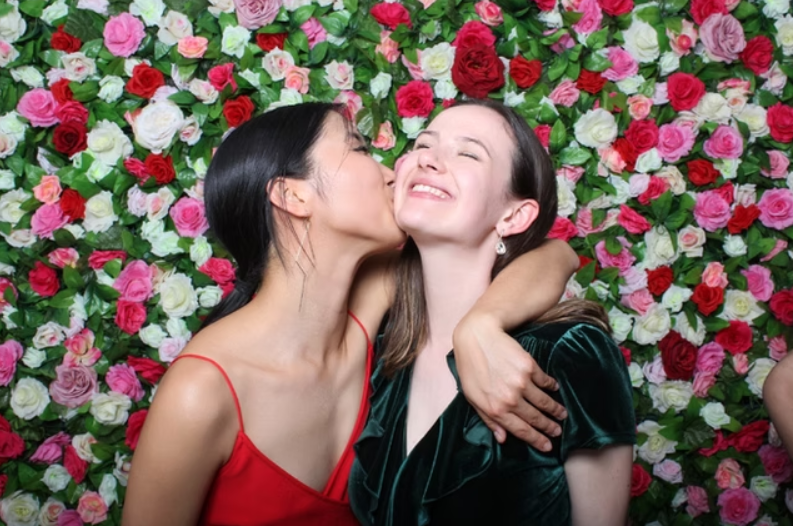 Two women are laughing and smiling at a party, holding drinks in front of a lush green floral backdrop with pink and white flowers, surrounded by other women.