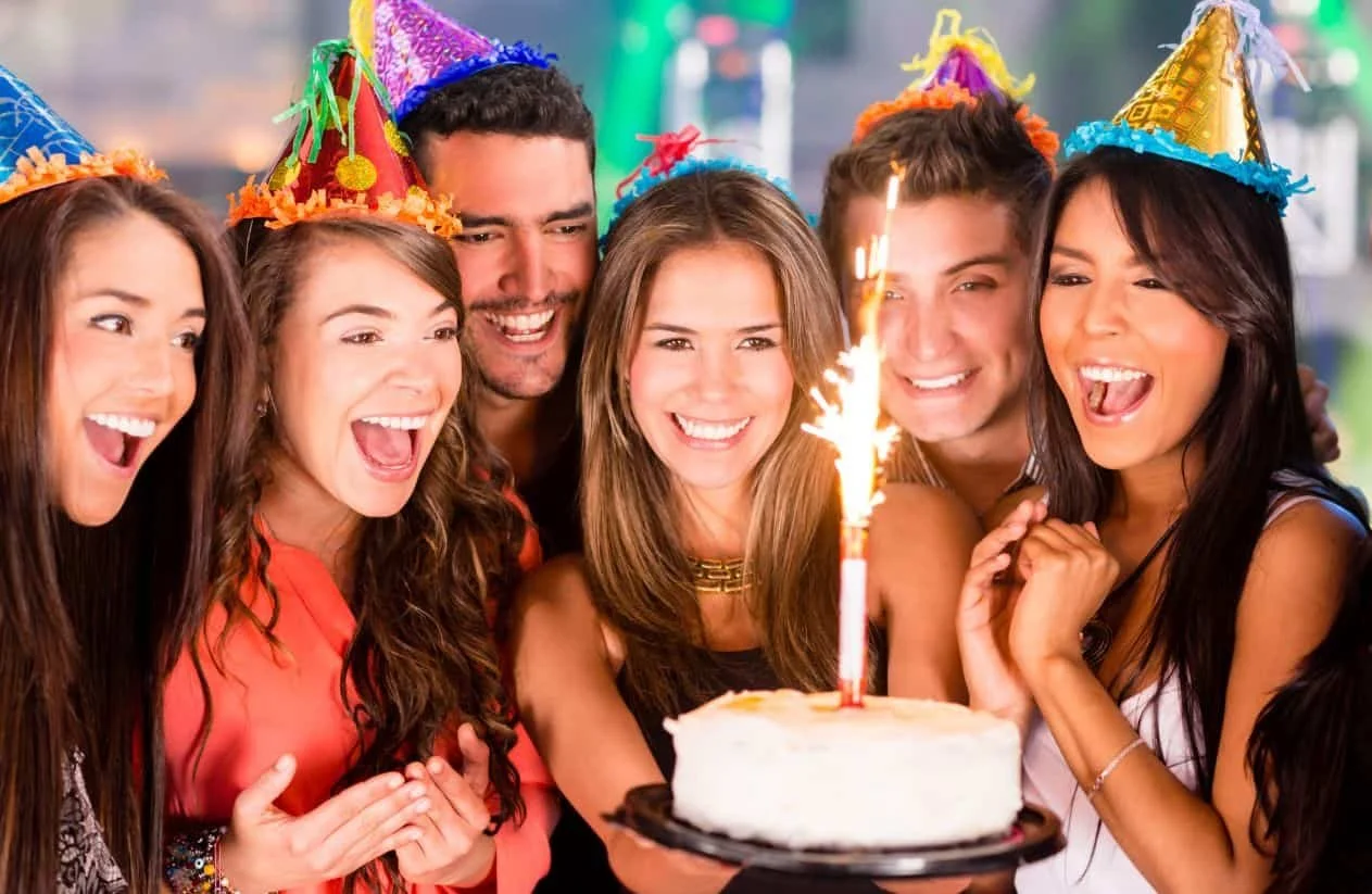 A group of six people celebrating a birthday, wearing party hats, gathered around a birthday cake with a lit sparkler, smiling and happy.