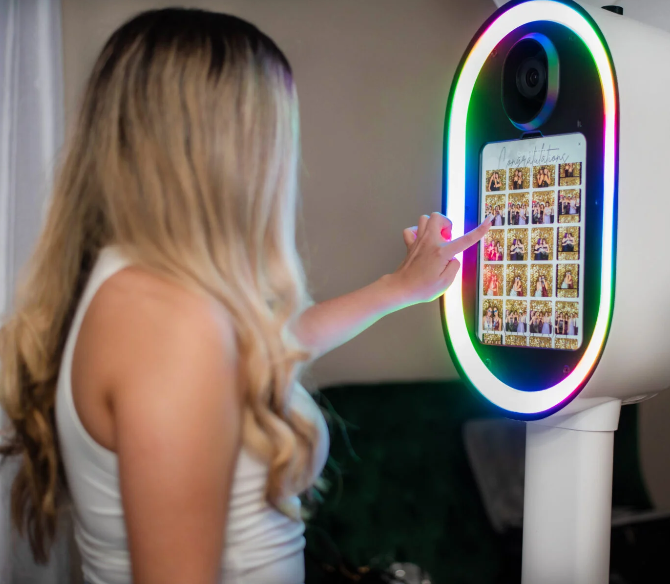 A young girl with long blonde hair in a white tank top interacts with a photo booth equipped with colorful LED lights and a touchscreen displaying a photo collage.