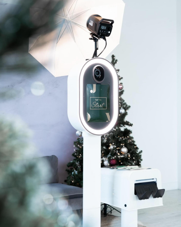 A festive holiday scene with a camera-based photo booth setup, including a camera, umbrella light, and a screen that says 'TAP TO Start', next to a decorated Christmas tree and a white bench.