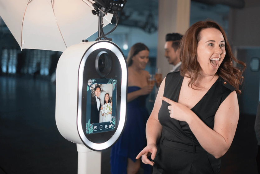 A woman with shoulder-length brown hair, wearing a black dress, is smiling and pointing at a photo booth with the screen showing a picture of a couple in formal attire. In the background, two people in blue dresses are chatting and holding drinks at what appears to be a party or event.