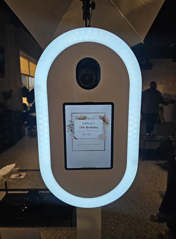 Photo booth with a camera and an illuminated oval frame displaying a screen that reads "Addison's 17th Birthday" with decorative plants and a rock graphic, at a birthday celebration.