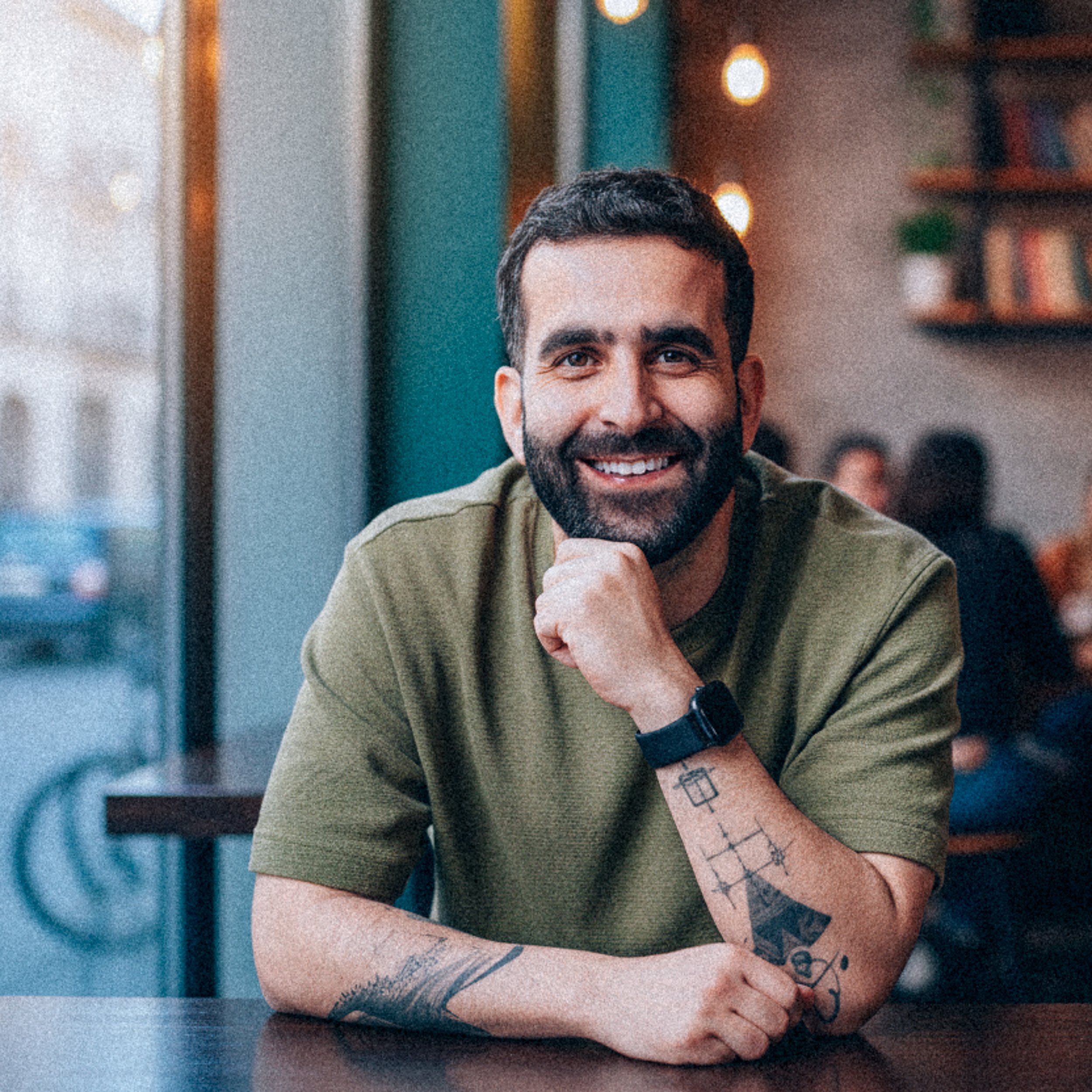 A smiling man with a beard and short dark hair, wearing a green t-shirt and a black watch, sitting at a table in a cozy cafe with warm lighting and bookshelves in the background.