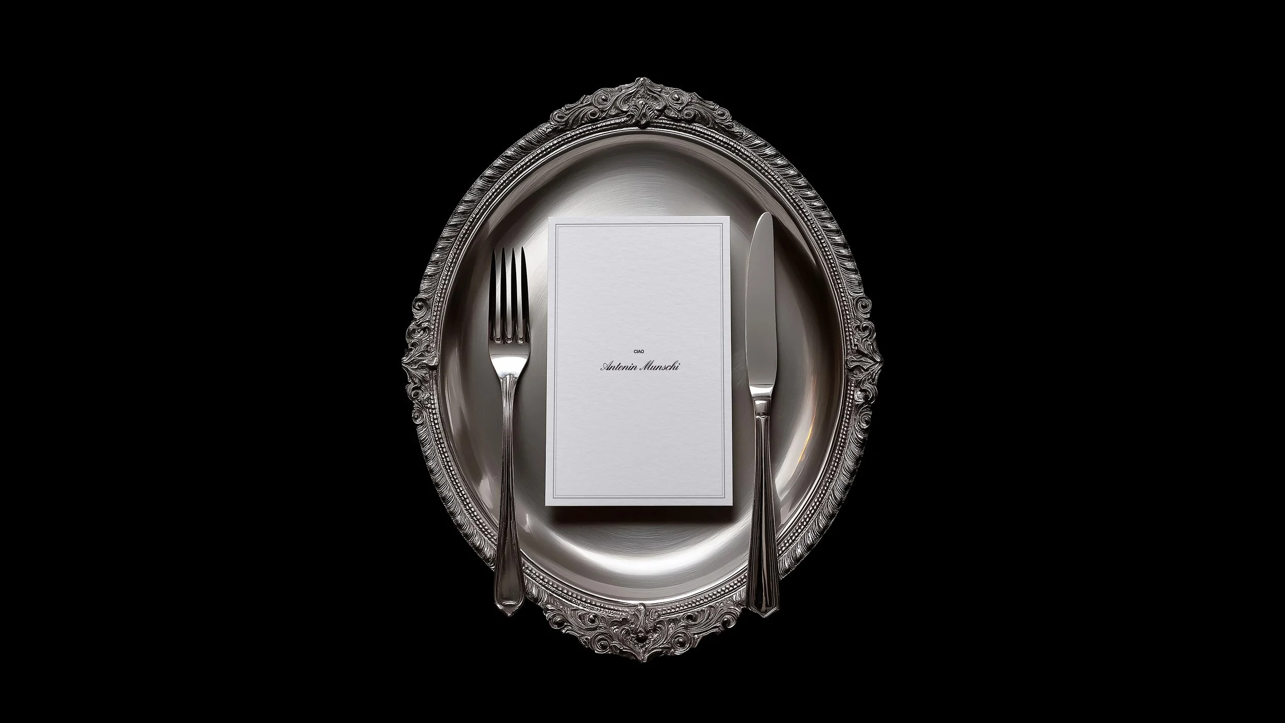 Elegant silver ornate plate with a fork and knife on either side of a napkin and menu card with the name Antonin Munschi on a black background.