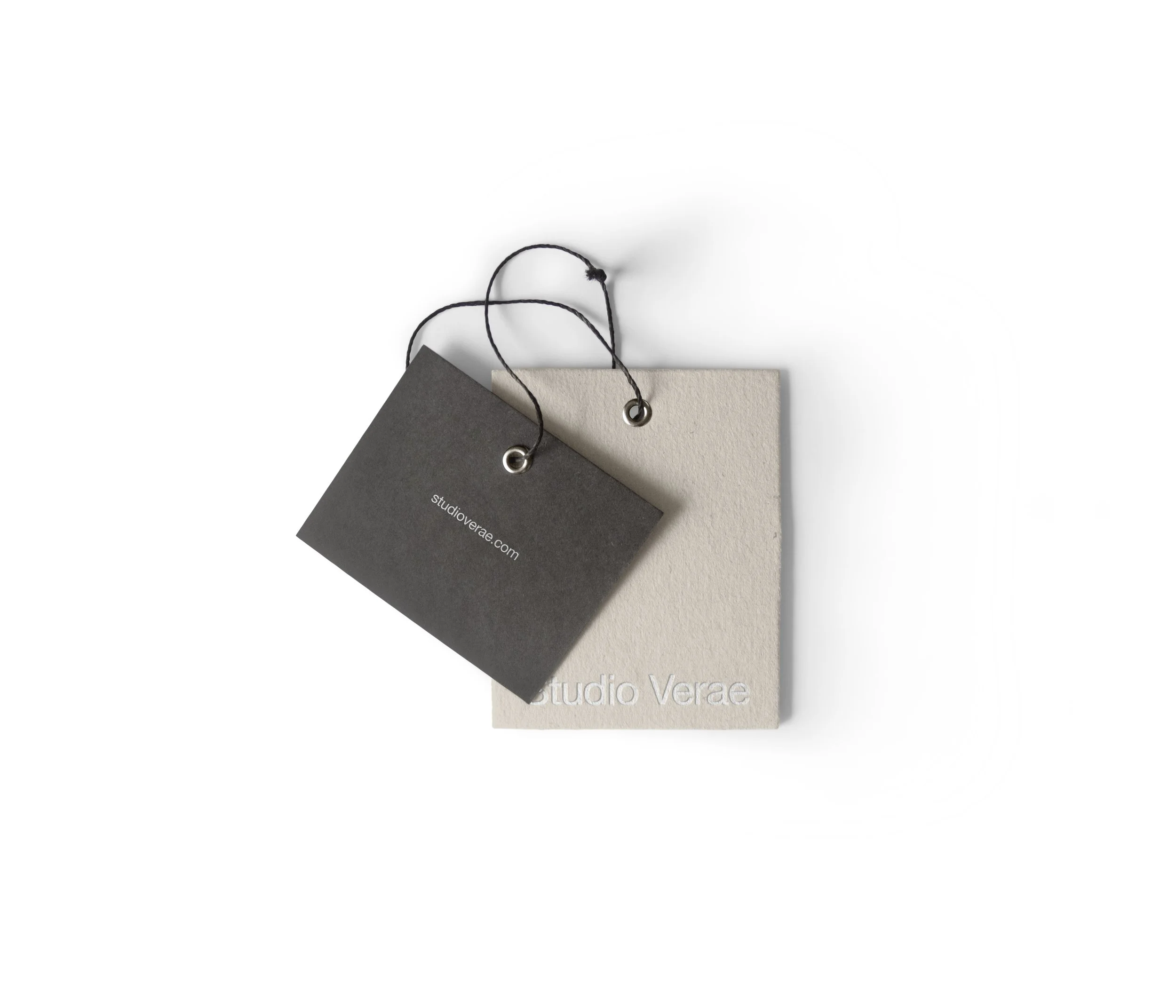 Two square paper tags, one dark gray and one light beige, with black string attached. The dark gray tag has the website 'studioverae.com' printed on it. The beige tag has 'Studio Verae' embossed in the bottom right corner.