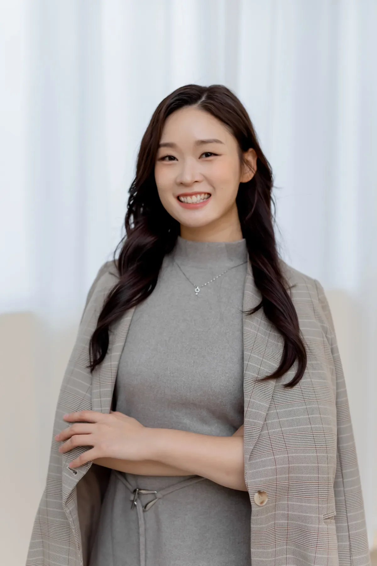Arienna Siew, Propnex Realtor, wearing a gray dress, a checkered blazer, and a delicate necklace, standing indoors in front of a white curtain.