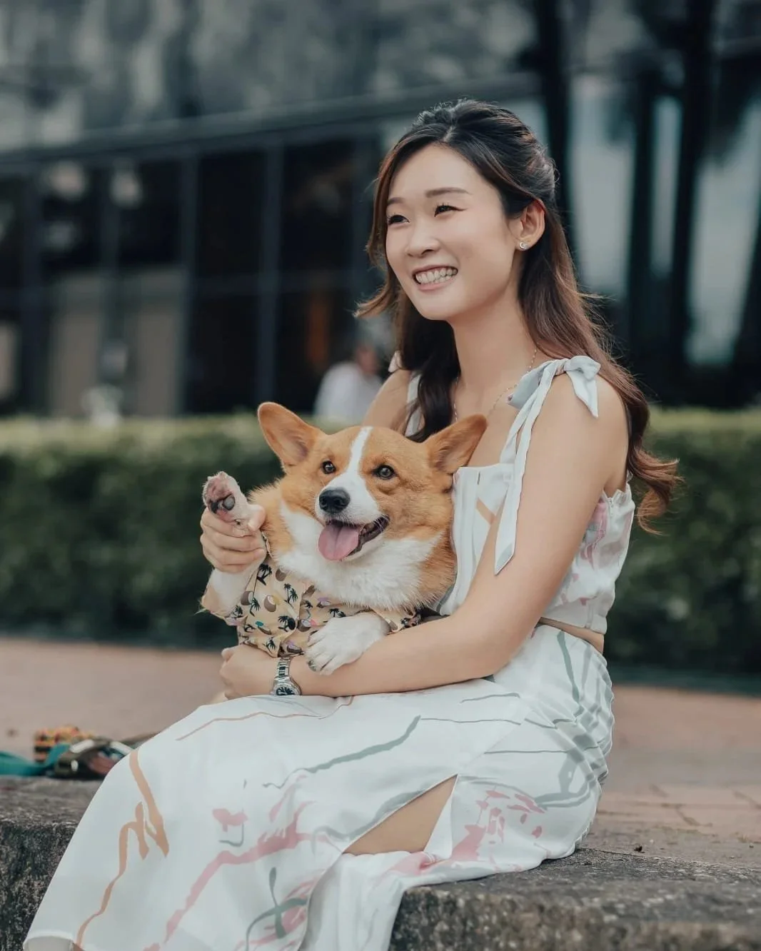 Adrienna sitting on a concrete ledge outdoors, smiling and holding a smiling corgi dog