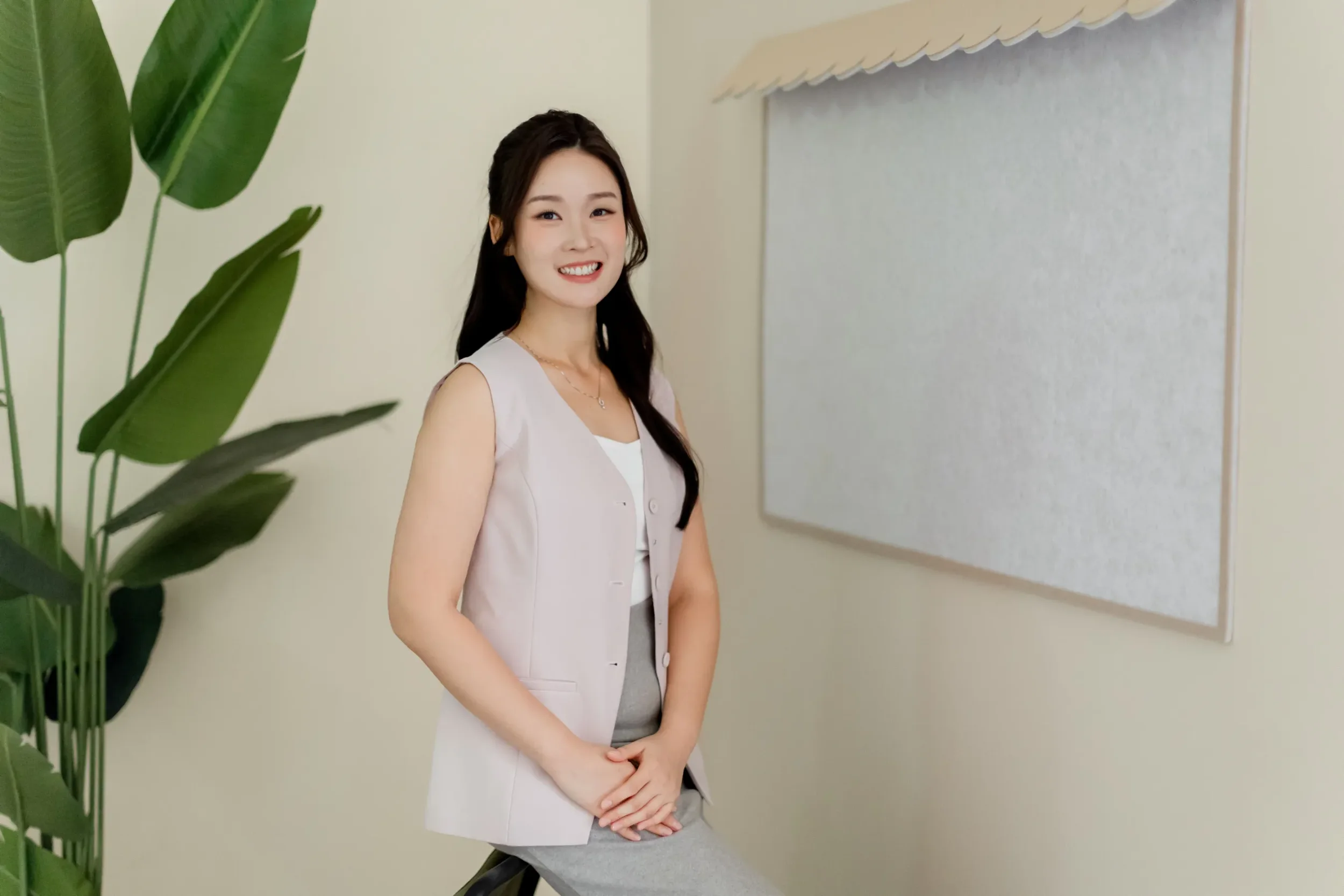 Adrienna Siew, Singapore Property Agent smiling, standing indoors near a leafy green plant and a blank beige bulletin board on a light-colored wall.