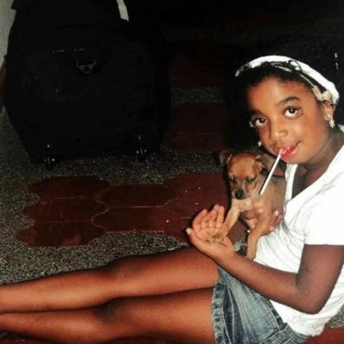 A young girl with a straw in her mouth, sitting on the floor holding a small dog. She is smiling and looking at the camera. There is a black suitcase behind her and a wooden table in front of her.