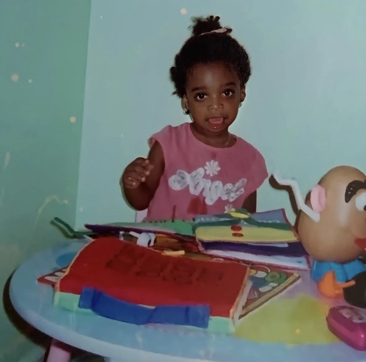 A young girl in a pink shirt sitting at a colorful table with toys, books, and a doll with a large head and painted face.
