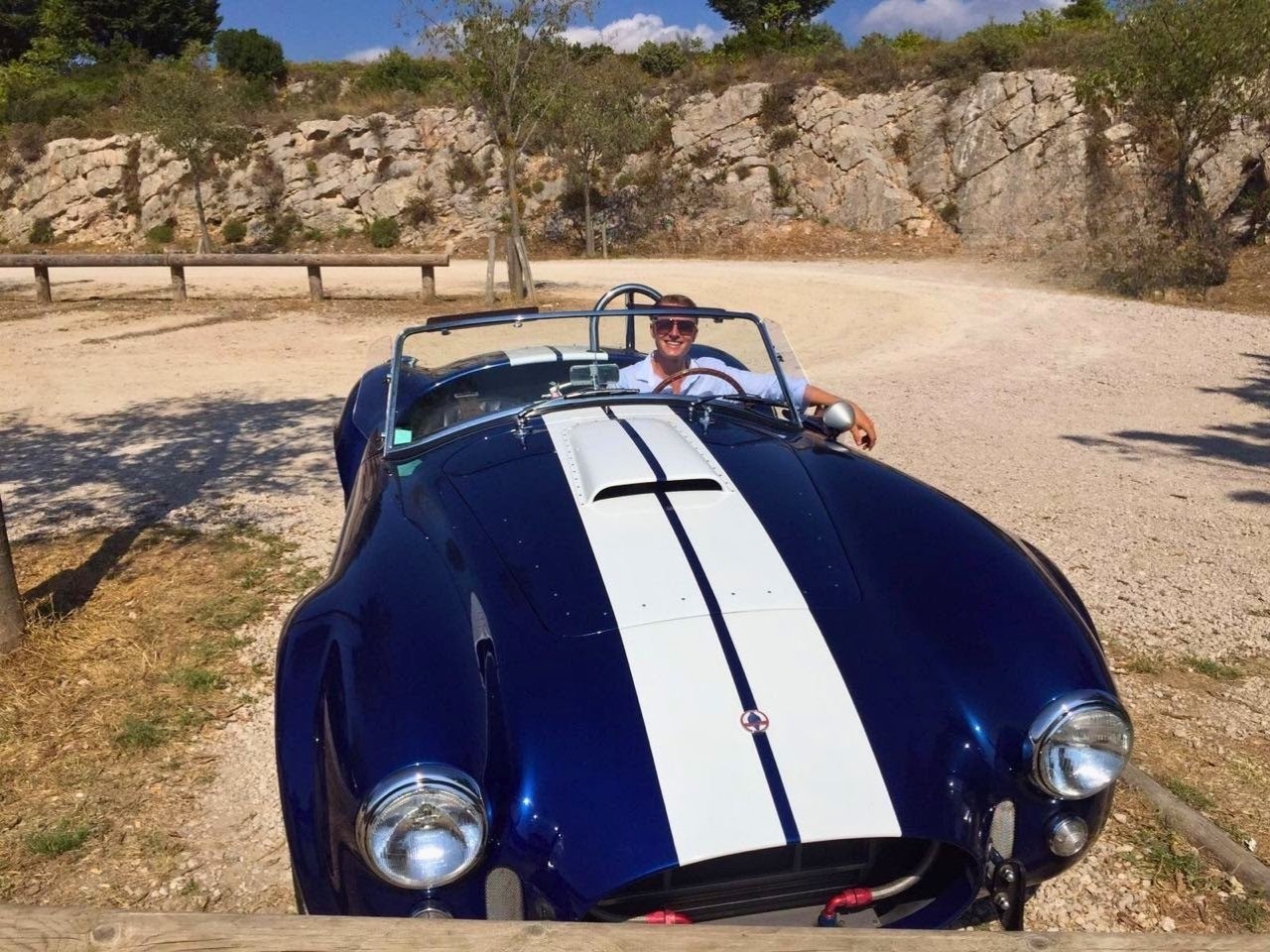 A person wearing sunglasses and a white shirt is sitting in a blue vintage convertible sports car with a white racing stripe, parked on a dirt area near trees and rocky terrain on a sunny day.
