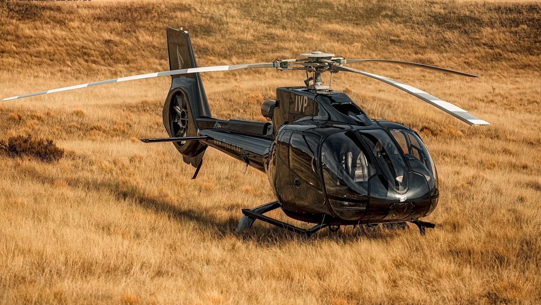 Black helicopter parked on a grassy field with dry, golden grass and rolling hills in the background.