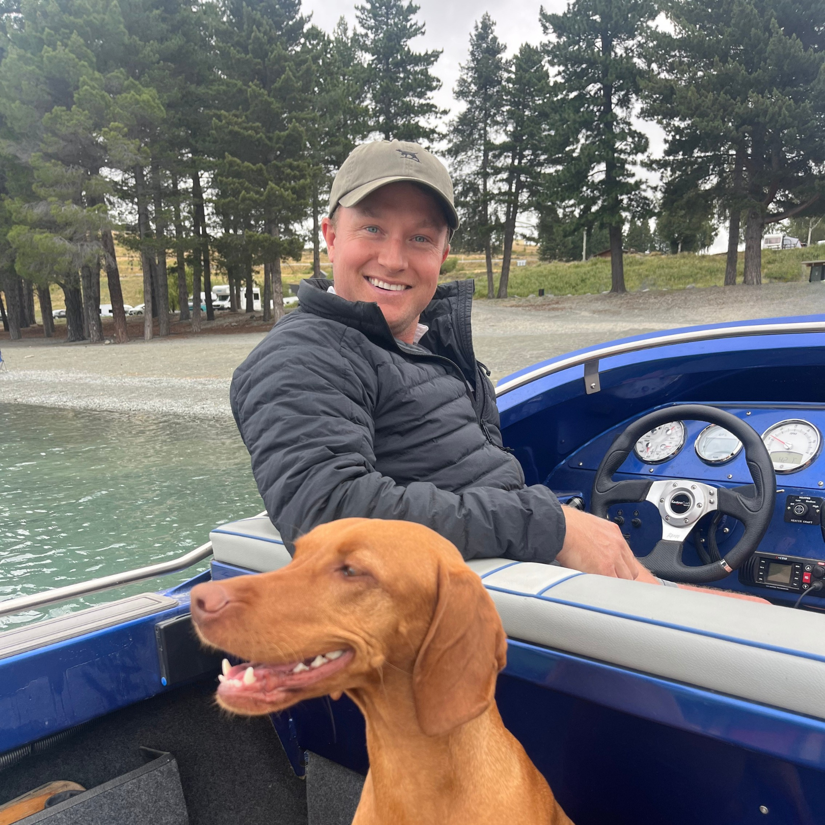 A smiling man in a beige cap and black jacket sitting in a blue boat on a lake. A brown dog with floppy ears is in the foreground, partially inside the boat. The background features trees along the shoreline and a cloudy sky.