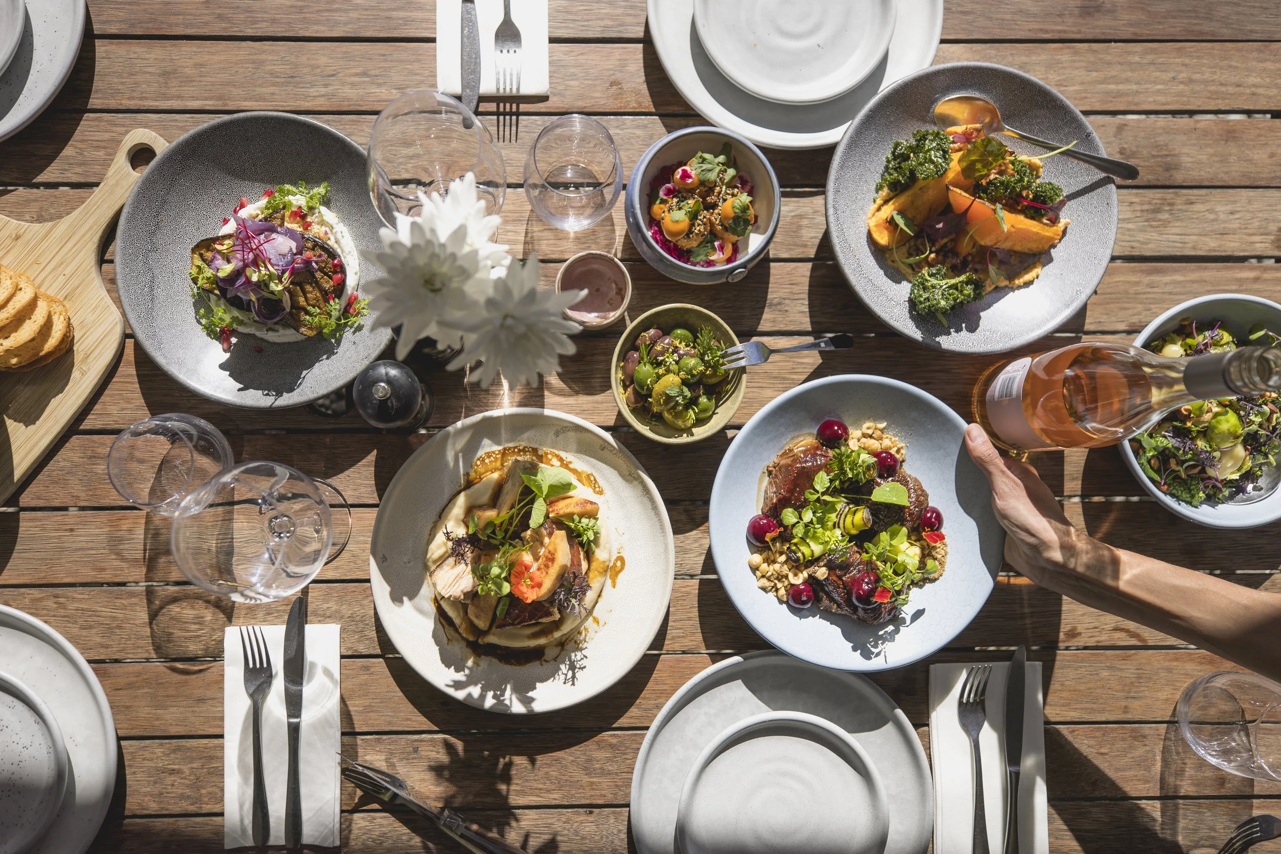 A wooden table set for a meal with various dishes including salads, bread, and drinks, with empty plates, glasses, and silverware arranged on napkins.