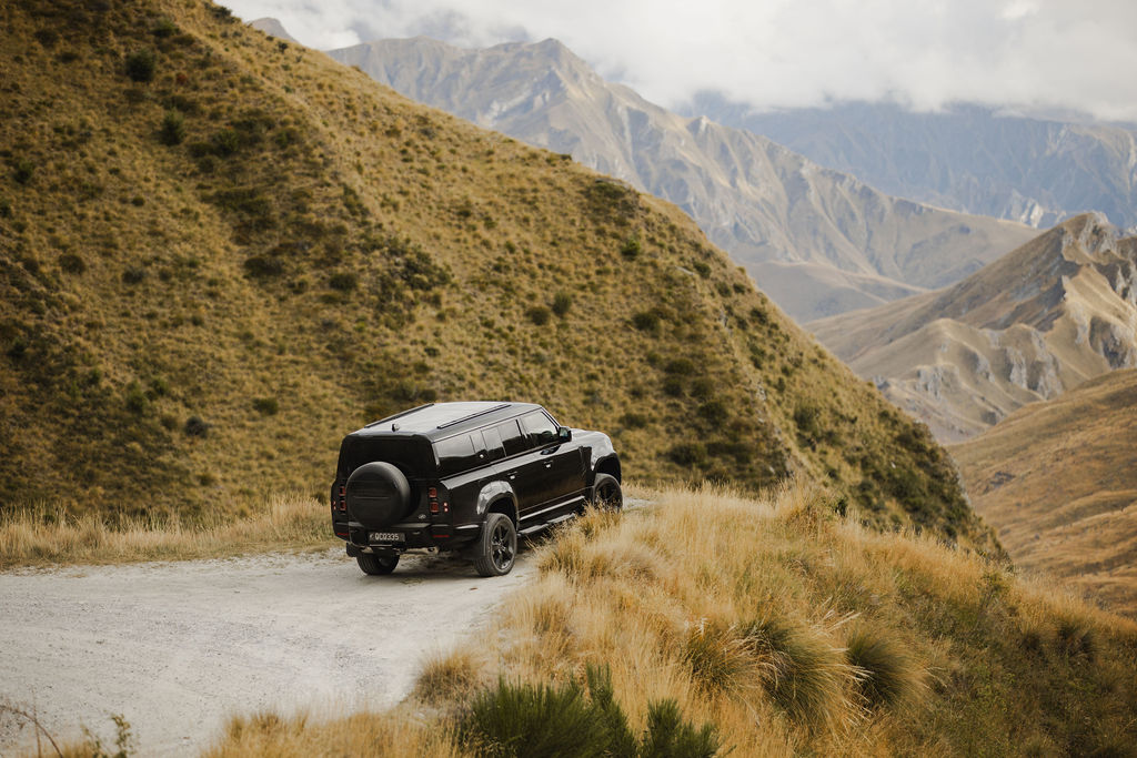 A black SUV parked on a gravel mountain road with rolling hills and rugged mountains in the background under a cloudy sky.