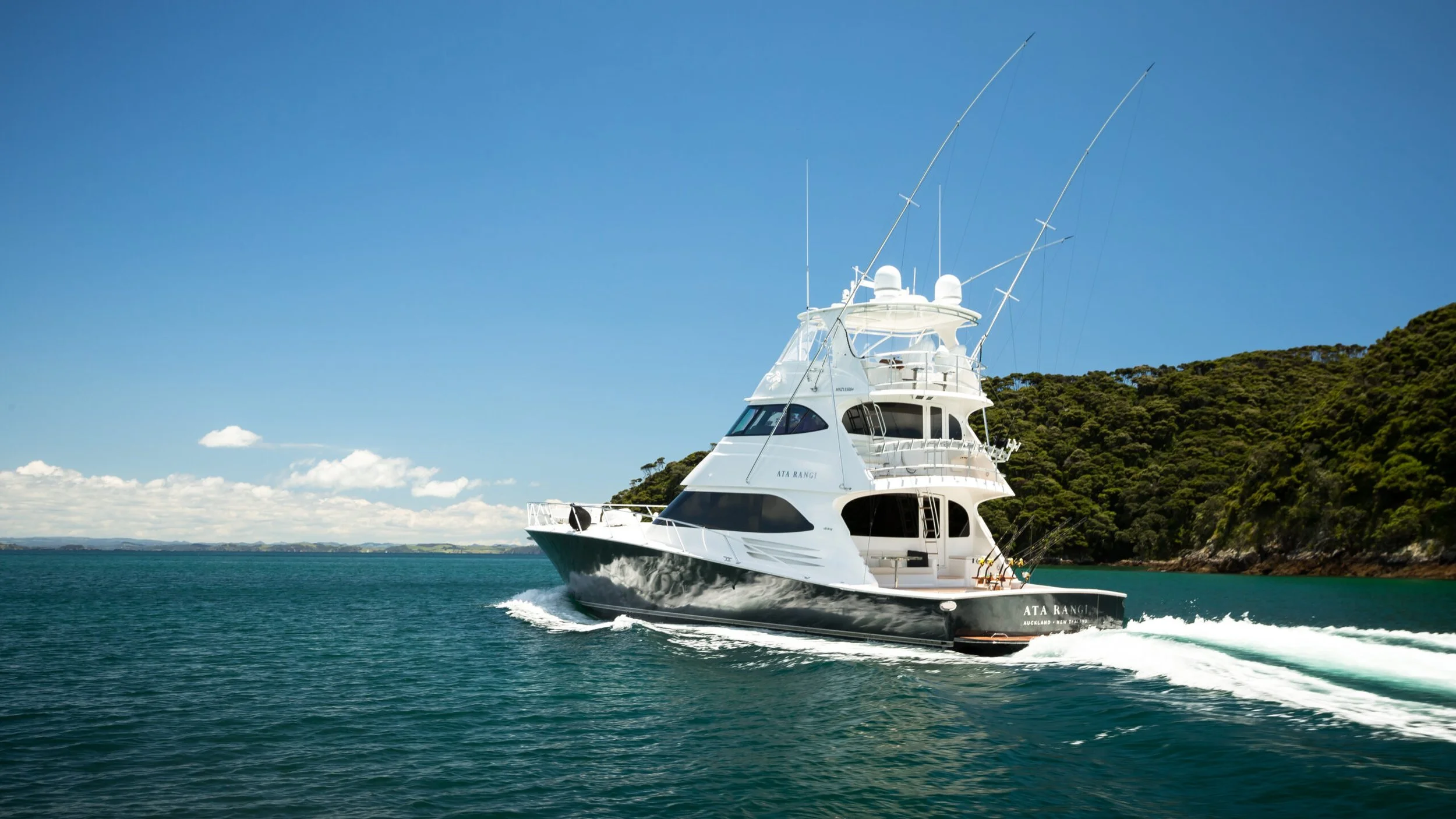 A white yacht named ATA RANGI cruising in a body of water near green land with trees, under a partly cloudy sky.