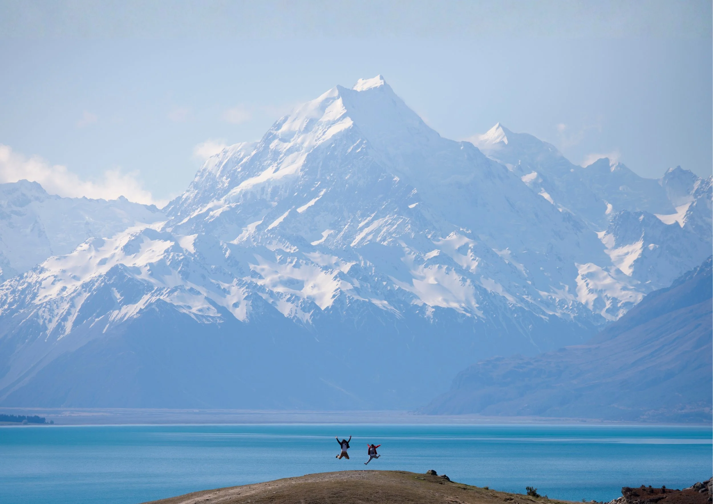 Two people jumping on a small hill overlooking a large body of water with a snow-capped mountain range in the background.