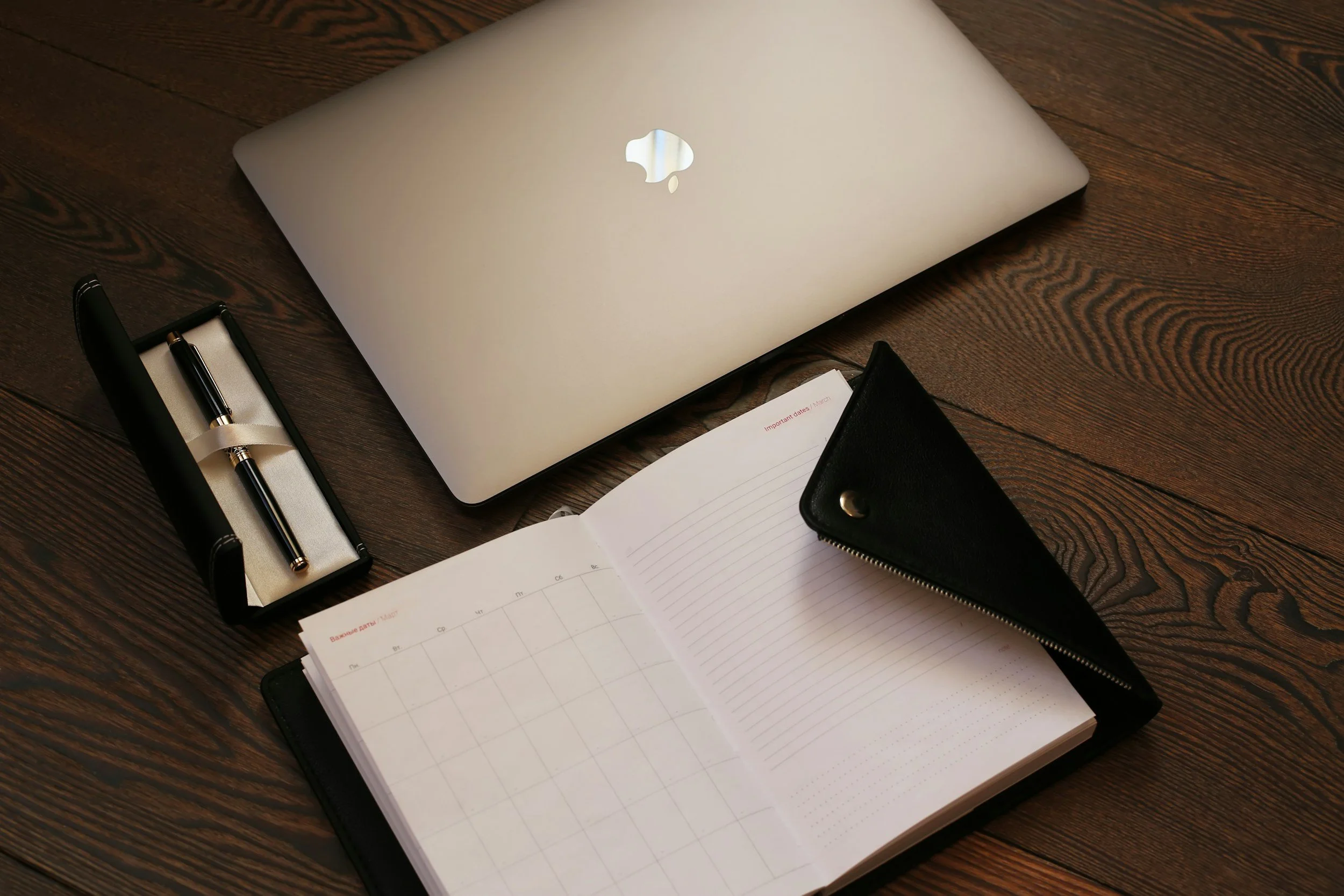 A laptop, pen in a case, planner, and a black zippered wallet on a wooden desk.