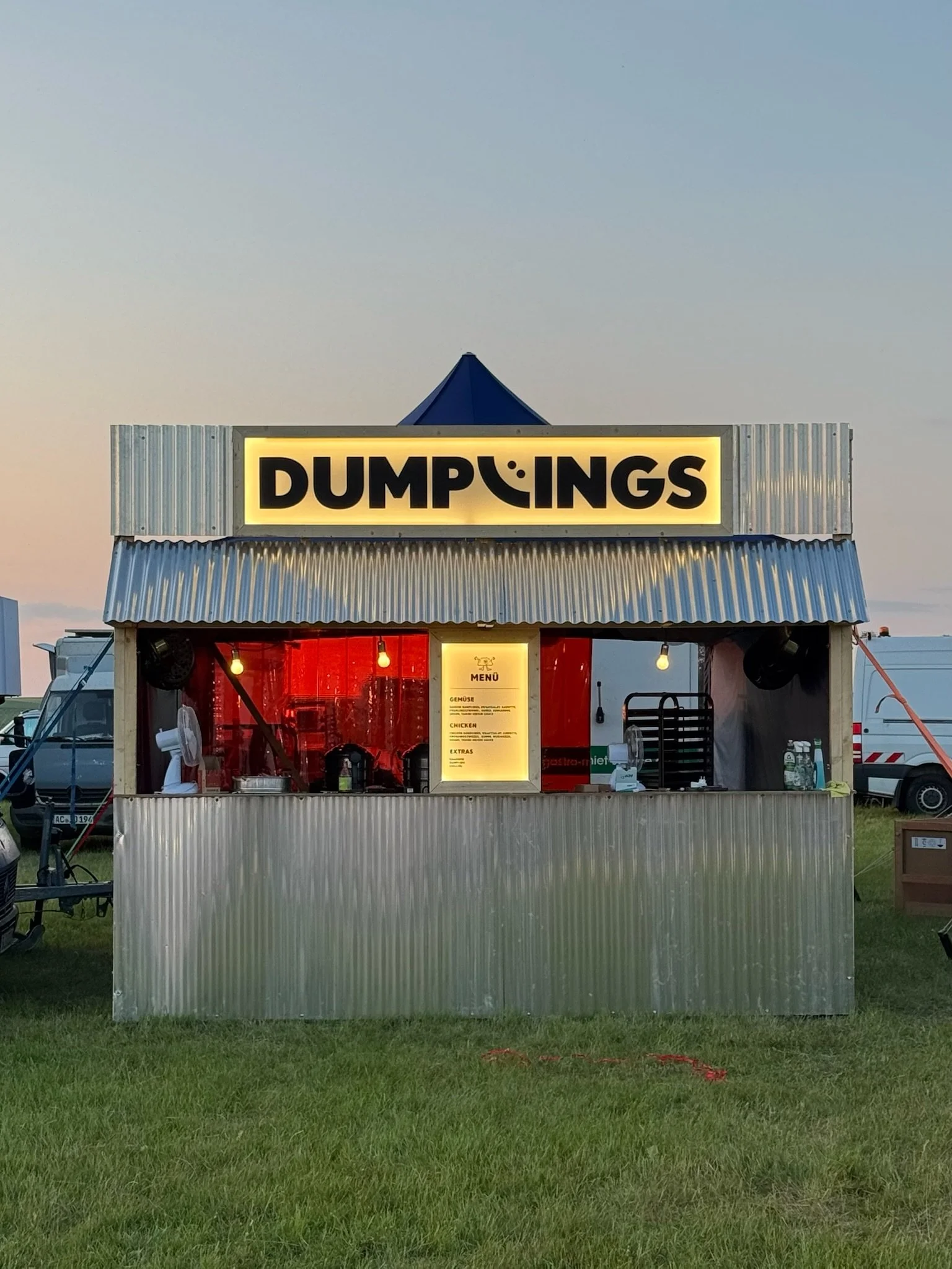 Ein Imbissstand aus Metall mit einem leuchtenden Schild, auf dem 'DUMPINGS' steht, befindet sich im Freien auf einer Wiese bei Sonnenuntergang.