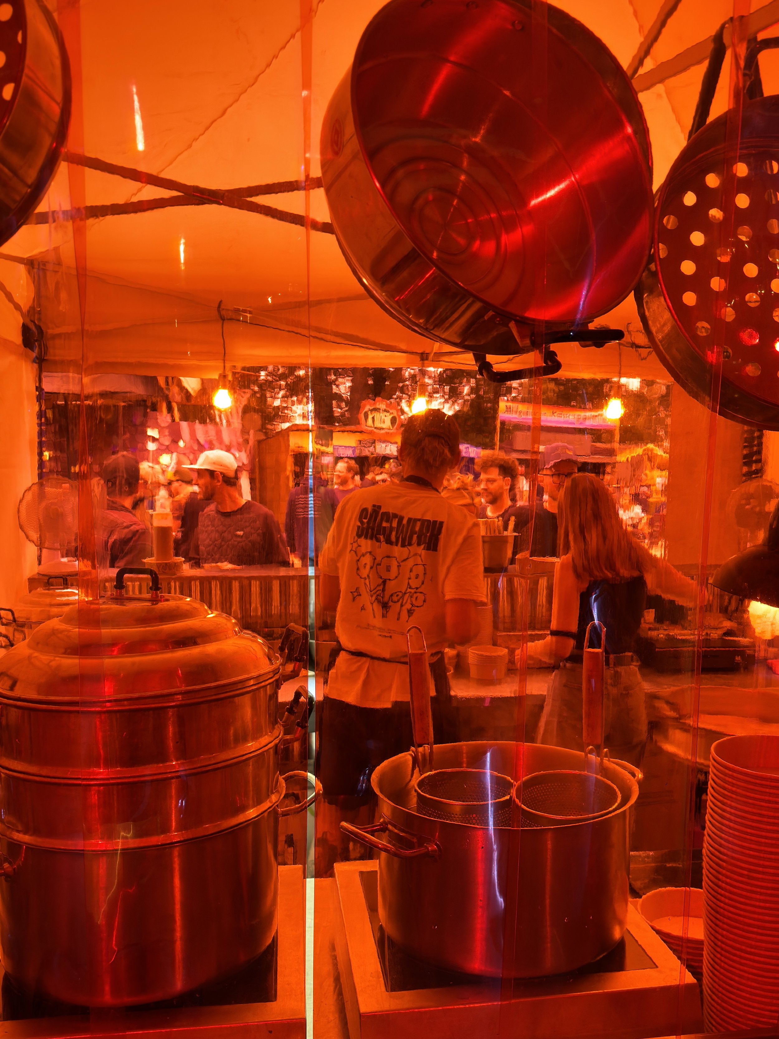 Blick durch eine Glaswand in einen Food-Festival- oder Marktstand mit zahlreichen Töpfen, Pfannen und Leuten im Hintergrund, umgeben von warmem, orangerotem Licht.