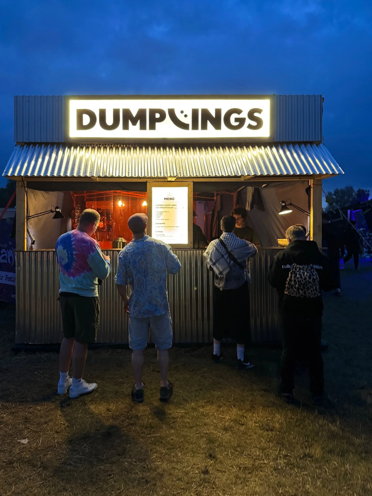 Menschen stehen vor einem Imbisswagen mit dem Schild 'DUMPINGS' in einer Nachtkulisse.