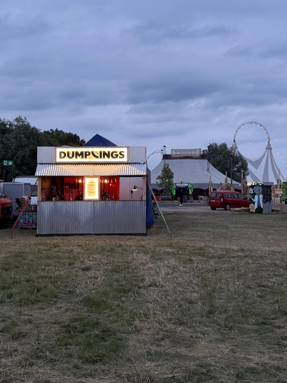 Ein fahrbarer Verkaufsstand mit einem leuchtenden Schild, das 'DUMPINGS' heißt, auf einem Jahrmarkt bei Dämmerung, im Hintergrund ein Riesenrad und Zelte.