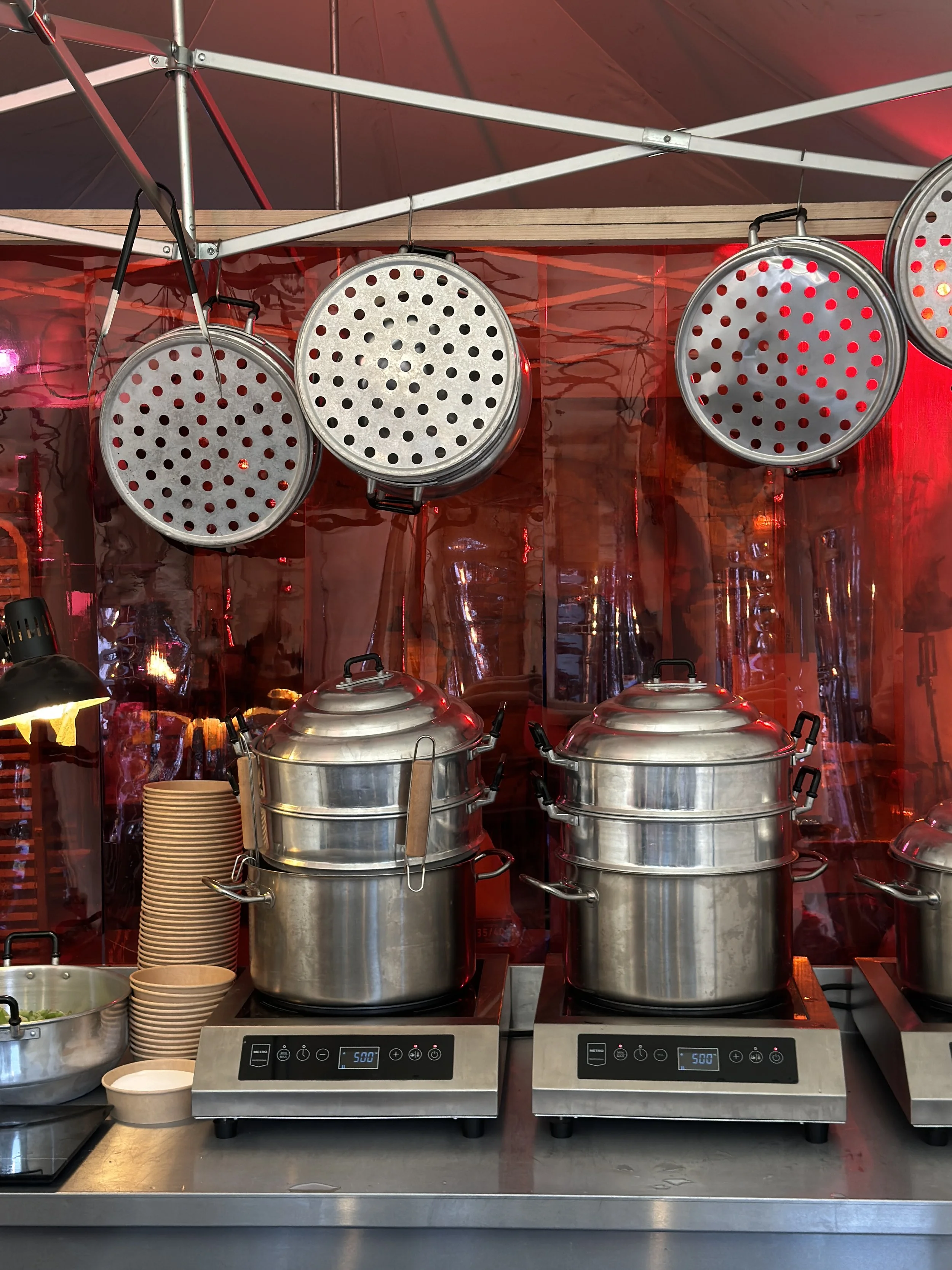 Zwei große Edelstahl-Dampfgarer auf einem Küchenbuffet, daneben sind Papier- und Suppenschalen. Über dem Buffet sind vier runde Edelstahl-Deckel zum Dampfgarer aufgehängt.