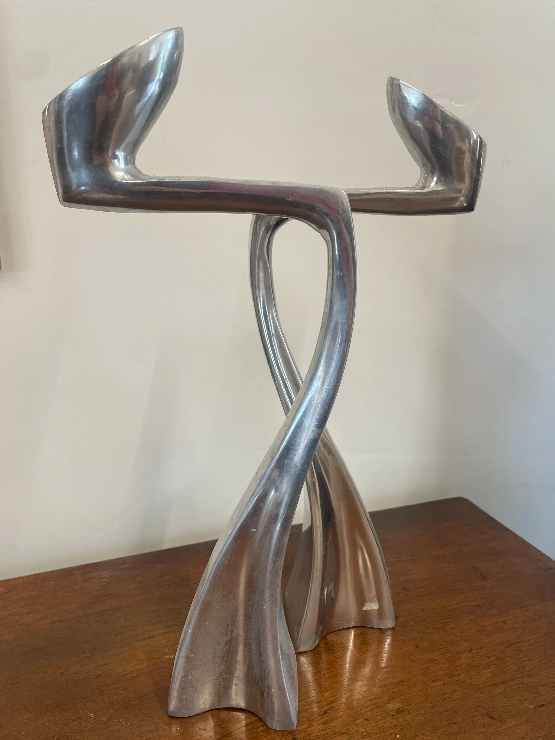 A modern abstract sculpture made of shiny metal with twisting, intertwined forms, placed on a wooden surface.