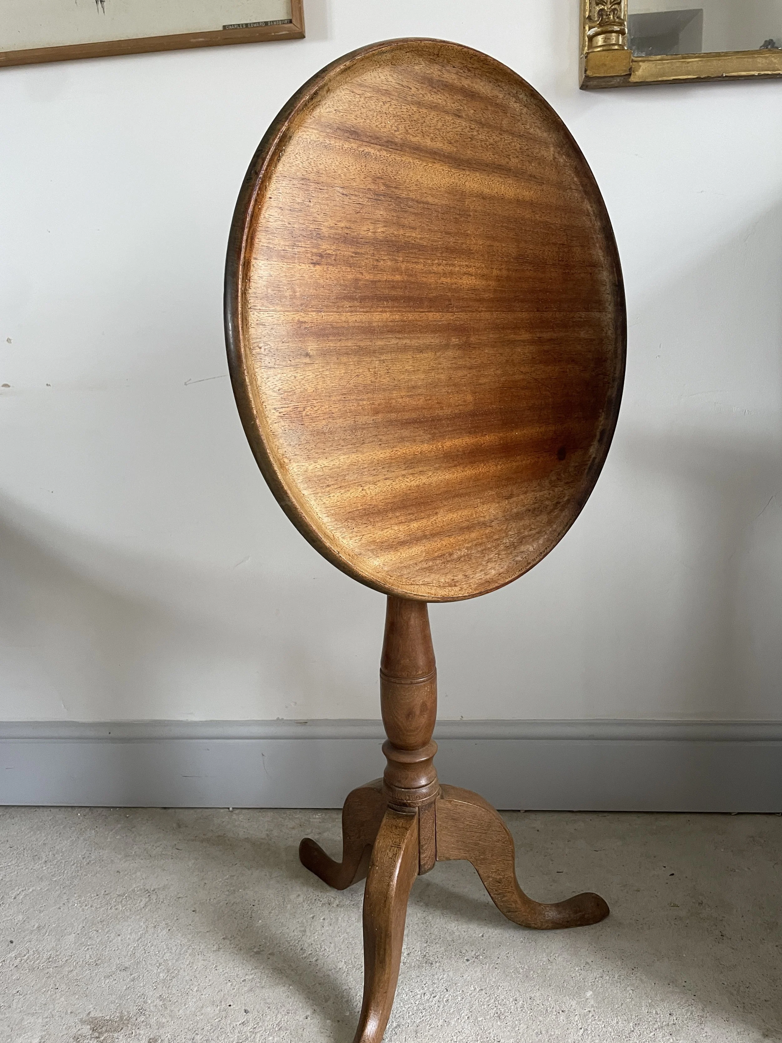 A round wooden table with a tilt, standing on a single turned pedestal with three curved legs on a carpeted floor against a white wall.
