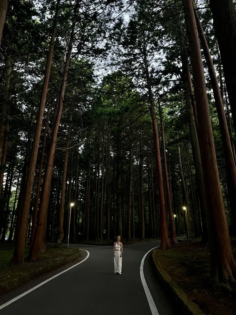 A woman standing on a winding forest road surrounded by tall trees.