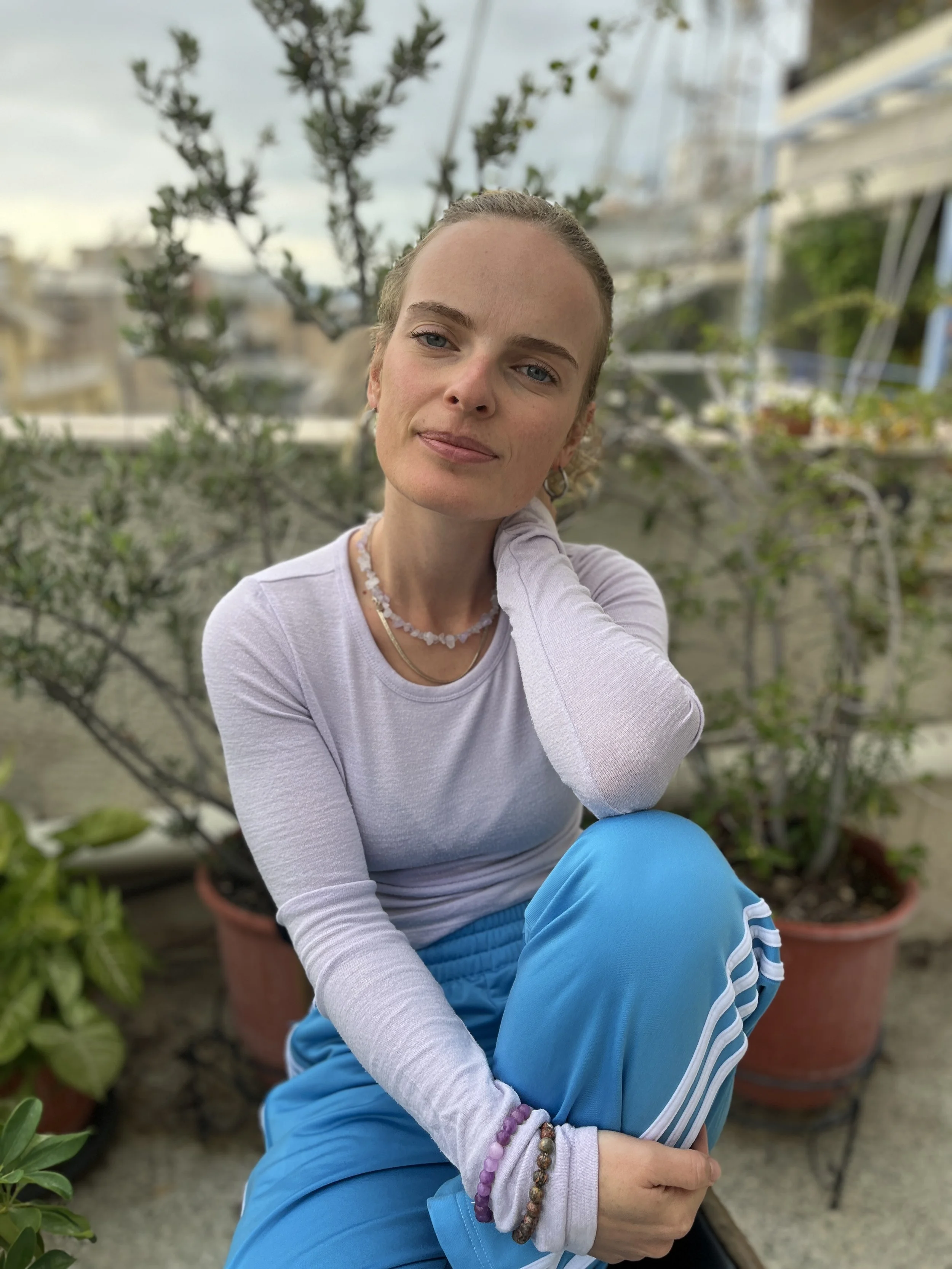 A woman with blonde hair tied back, wearing a long-sleeve white shirt, blue athletic pants, and several bracelets, sitting outdoors on a rooftop garden, posing with her head tilted slightly and a relaxed expression, with potted plants and a cityscape in the background.
