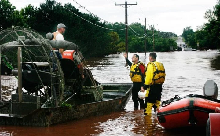 Rescue workers in yellow life jackets assist a person onto a boat during a flood, with water covering the street and power lines overhead.